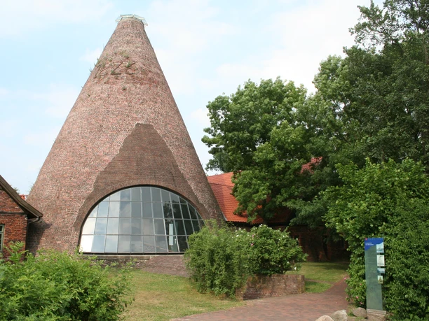 Glashütte Gernheim Historische glasfabriek met opvallende bakstenen toren en glazen façade, omgeven door groene vegetatie.