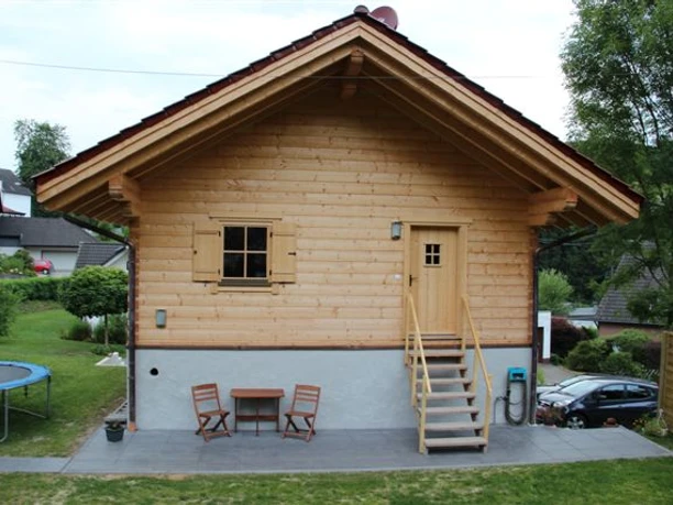 Ferienhaus Breibach Kleines Holzhaus mit Satteldach, kleiner Terrasse, Tisch und Stühlen, umgeben von grüner Landschaft.