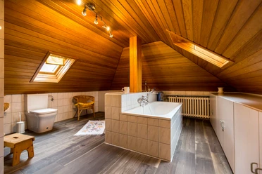 Mediendatei Dachgeschoss-Badezimmer mit Holzverkleidung, Dachfenster, Badewanne, Toilette und Sessel.