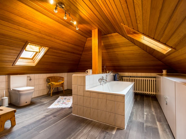 Mediendatei Dachgeschoss-Badezimmer mit Holzverkleidung, Dachfenster, Badewanne, Toilette und Sessel.
