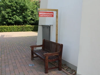 Eine hölzerne Mitfahrerbank steht auf einem gepflasterten Platz in der Nähe einer Windmühle.