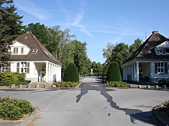 Eingang zum Friedhof Zwei weiße, symmetrische Gebäude flankieren den Zugang zu einem Friedhof, umgeben von Bäumen.