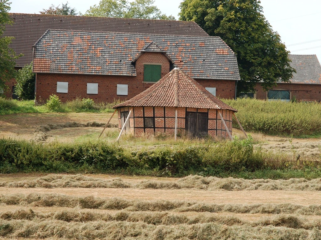 Ein historisches Göpelgebäude mit Holzfassade und Ziegeldach, umgeben von Feldern und einem Bauernhaus.