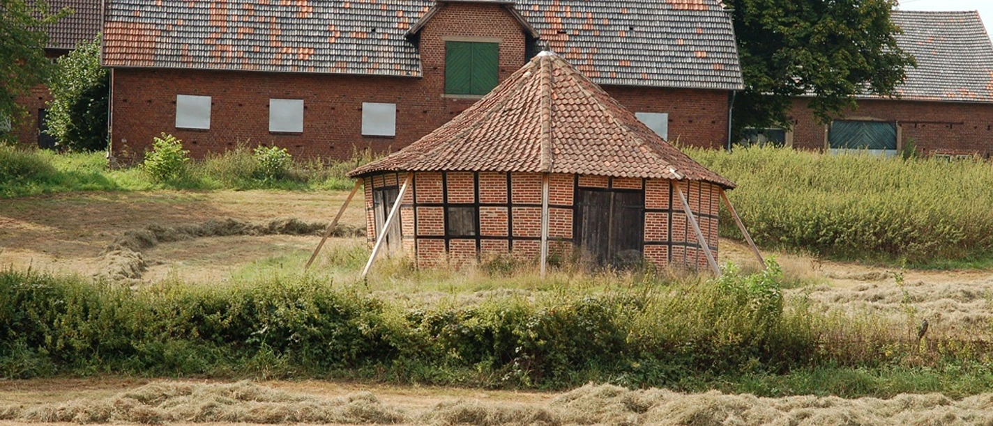 Ein historisches Göpelgebäude mit Holzfassade und Ziegeldach, umgeben von Feldern und einem Bauernhaus.