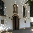 Eingang der weißen Pfarrkirche St. Joseph in Paderborn-Marienloh mit brauner Holztür unter Bäumen.