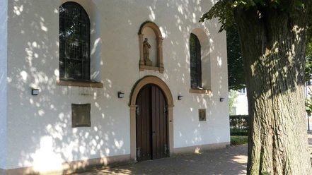 Eingang der Pfarrkirche St. Joseph in Paderborn-Marienloh Eingang der weißen Pfarrkirche St. Joseph in Paderborn-Marienloh mit brauner Holztür unter Bäumen.