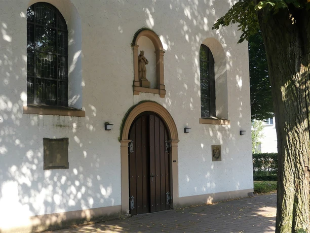 Eingang der Pfarrkirche St. Joseph in Paderborn-Marienloh Eingang der weißen Pfarrkirche St. Joseph in Paderborn-Marienloh mit brauner Holztür unter Bäumen.