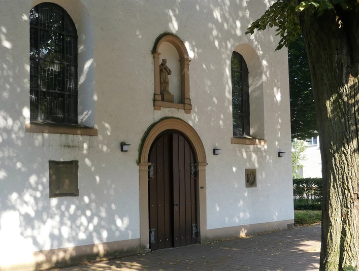 Eingang der Pfarrkirche St. Joseph in Paderborn-Marienloh Eingang der weißen Pfarrkirche St. Joseph in Paderborn-Marienloh mit brauner Holztür unter Bäumen.