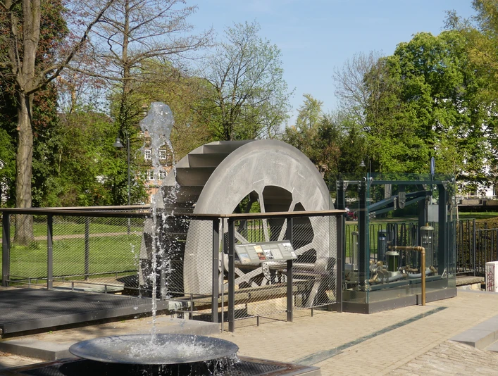Ein Wasserrad-Modell in einem Park, umgeben von Bäumen und einer sprudelnden Fontäne.