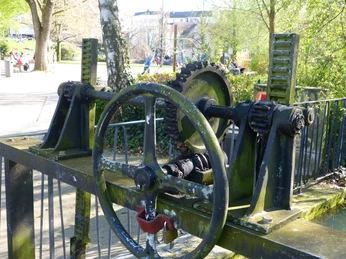 Mechanisches Wasserrad und Zahnräder aus Metall, umgeben von Frühlingsbäumen und Spaziergängern.