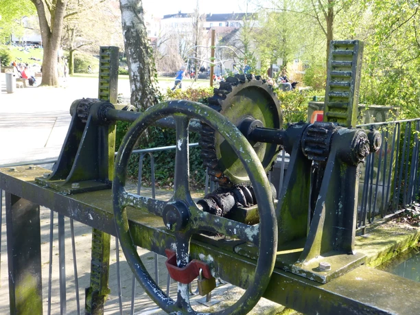 Wehr beim Funktionsmodell der Wasserkunst Mechanisches Wasserrad und Zahnräder aus Metall, umgeben von Frühlingsbäumen und Spaziergängern.