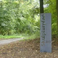 Stahlstele mit der Aufschrift "Skulpturenpfad 2013" vor einem bewaldeten Wegesrand.