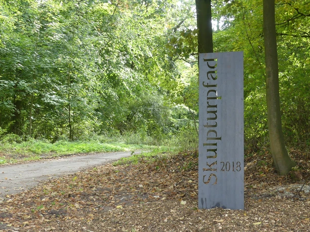 Stahlstele mit der Aufschrift "Skulpturenpfad 2013" vor einem bewaldeten Wegesrand.