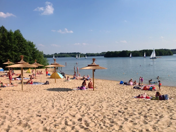 Sandstrand am Nordufer beim "Eisklang"-Bistro Sandstrand mit Badenden und Segelbooten auf einem See, umgeben von grünen Bäumen im Hintergrund.