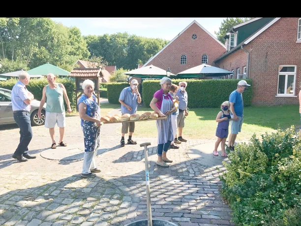 Backtag an der Hengstforder Mühle Eine Gruppe von Menschen in sommerlicher Kleidung und Hüten genießt den sonnigen Tag und betrachtet frisch gebackene Brote auf einer Platte vor der Hengstforder Mühle. Die Szenerie ist geprägt von Fröhlichkeit und Bewegung.