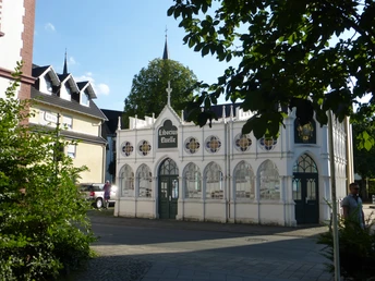 Historische Liborius-Trinkhalle in Bad Lippspringe vor einer Kirche, umgeben von Laubbäumen.