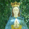Marienstatue mit goldener Krone und betenden Händen, umgeben von grünem Blätterwerk im Haxtergrund.