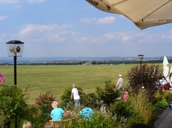 Blick von einer Terrasse auf eine weite Wiesenlandschaft mit Menschen und Sonnenschirmen.