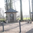 Steinerne Kapelle mit metallenen Toren, umgeben von Bäumen und Bänken auf einem kleinen Platz.
