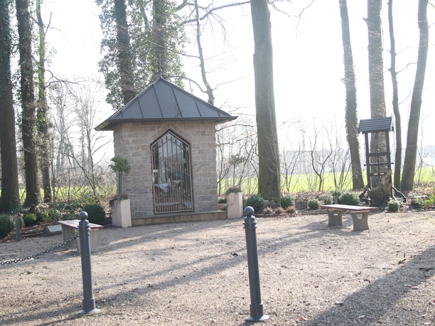 Steinerne Kapelle mit metallenen Toren, umgeben von Bäumen und Bänken auf einem kleinen Platz.