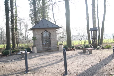 Kapelle in Sende Steinerne Kapelle mit metallenen Toren, umgeben von Bäumen und Bänken auf einem kleinen Platz.