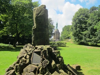 Großer, markanter Stein im Vordergrund, mit Hermannsdenkmal im Hintergrund, umgeben von Wald.