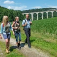 Drei Wanderer genießen einen Spaziergang auf einem Feldweg, mit einem Zug auf einem Viadukt im Hintergrund.
