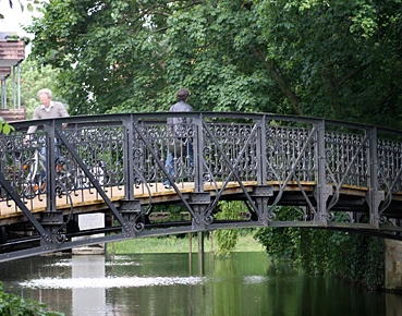 Herthabrücke Fußgängerbrücke mit schmiedeeisernem Geländer, umgeben von üppigem Grün, überquert einen Fluss.
