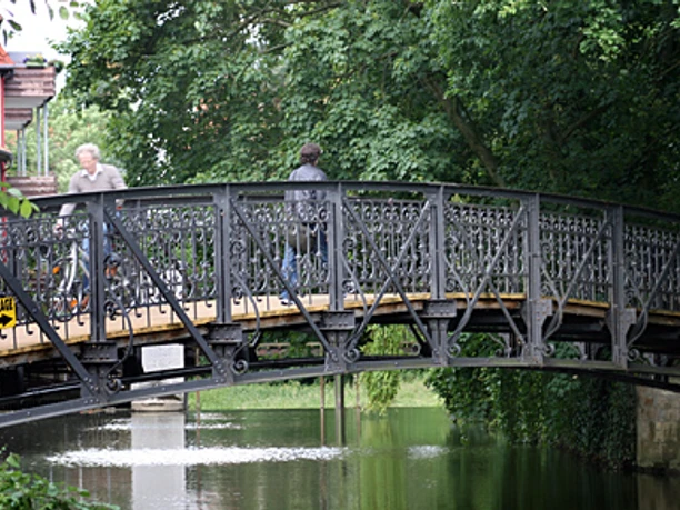 Herthabrücke Fußgängerbrücke mit schmiedeeisernem Geländer, umgeben von üppigem Grün, überquert einen Fluss.
