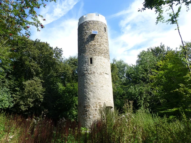Ein runder Steinturm erhebt sich inmitten eines grünen Waldes unter einem blauen Himmel mit Wolken.