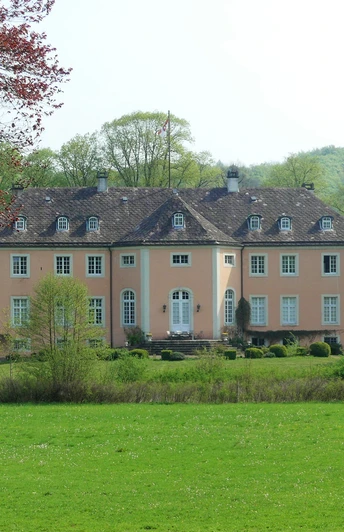 Schloss Rheder von der Gartenseite, umgeben von grünen Wiesen und Bäumen, mit sandfarbenem Mauerwerk.