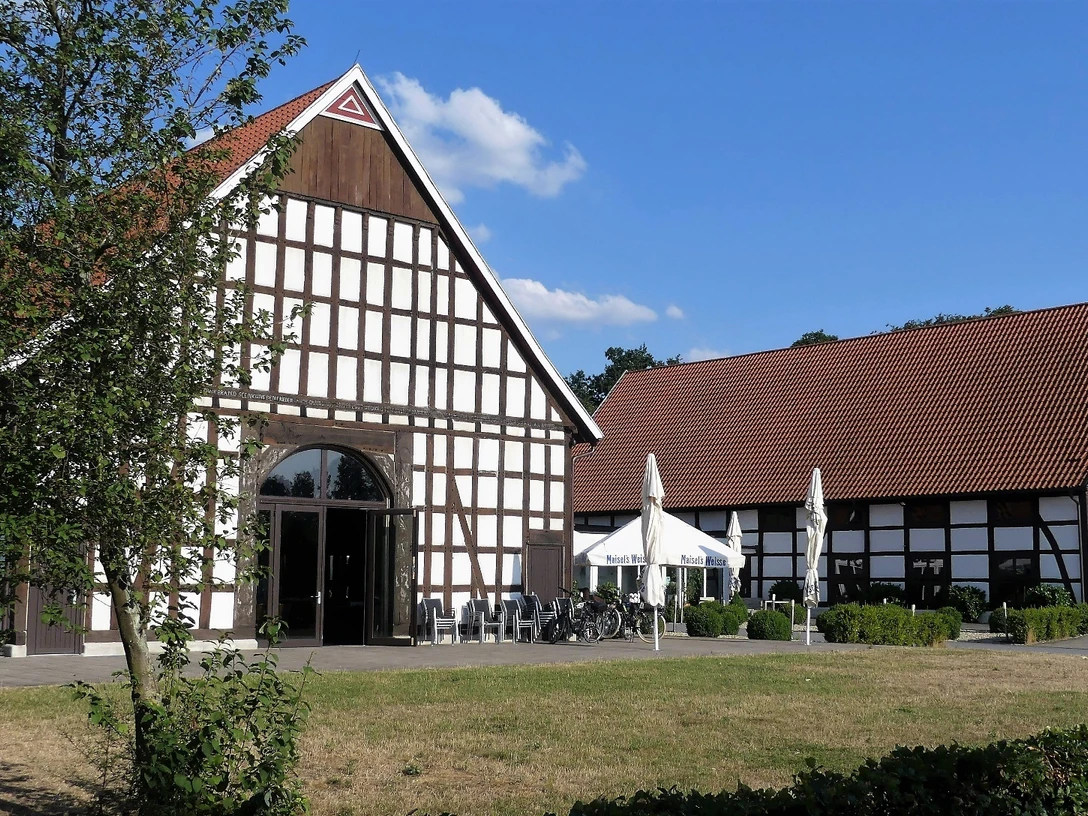 Ein Fachwerkgebäude mit roten Ziegeldächern und Terrasse, im Vordergrund Bäume und Rasenfläche.