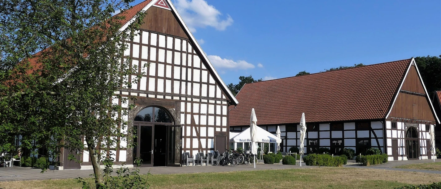 Ein Fachwerkgebäude mit roten Ziegeldächern und Terrasse, im Vordergrund Bäume und Rasenfläche.