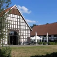 Gut Lippesee Ein Fachwerkgebäude mit roten Ziegeldächern und Terrasse, im Vordergrund Bäume und Rasenfläche.