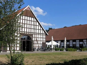 Gut Lippesee Ein Fachwerkgebäude mit roten Ziegeldächern und Terrasse, im Vordergrund Bäume und Rasenfläche.