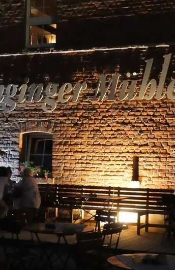 Die Alt-Enginger MĂĽhle beleuchtet, mit Besuchern im stimmungsvollen AuĂźenbereich am Abend.