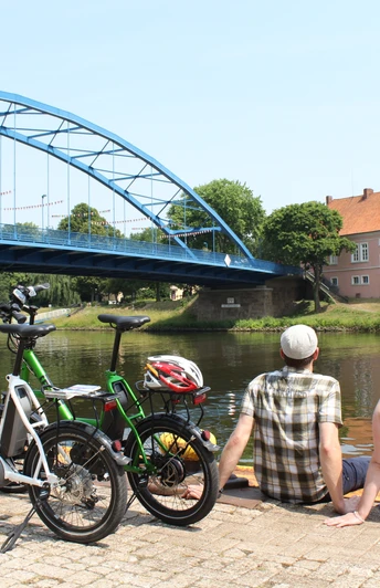 Ein Paar sitzt entspannt am Wasser, mit Blick auf das historische Grafenschloss zu Hoya und eine blaue Brücke.A couple sits relaxed by the water, with a view of the historic Hoya Castle and a blue bridge.Et par sidder afslappet ved vandet med udsigt til det historiske Hoya-slot og en blå bro.Een koppel zit ontspannen aan het water, met uitzicht op het historische Hoya Kasteel en een blauwe brug.