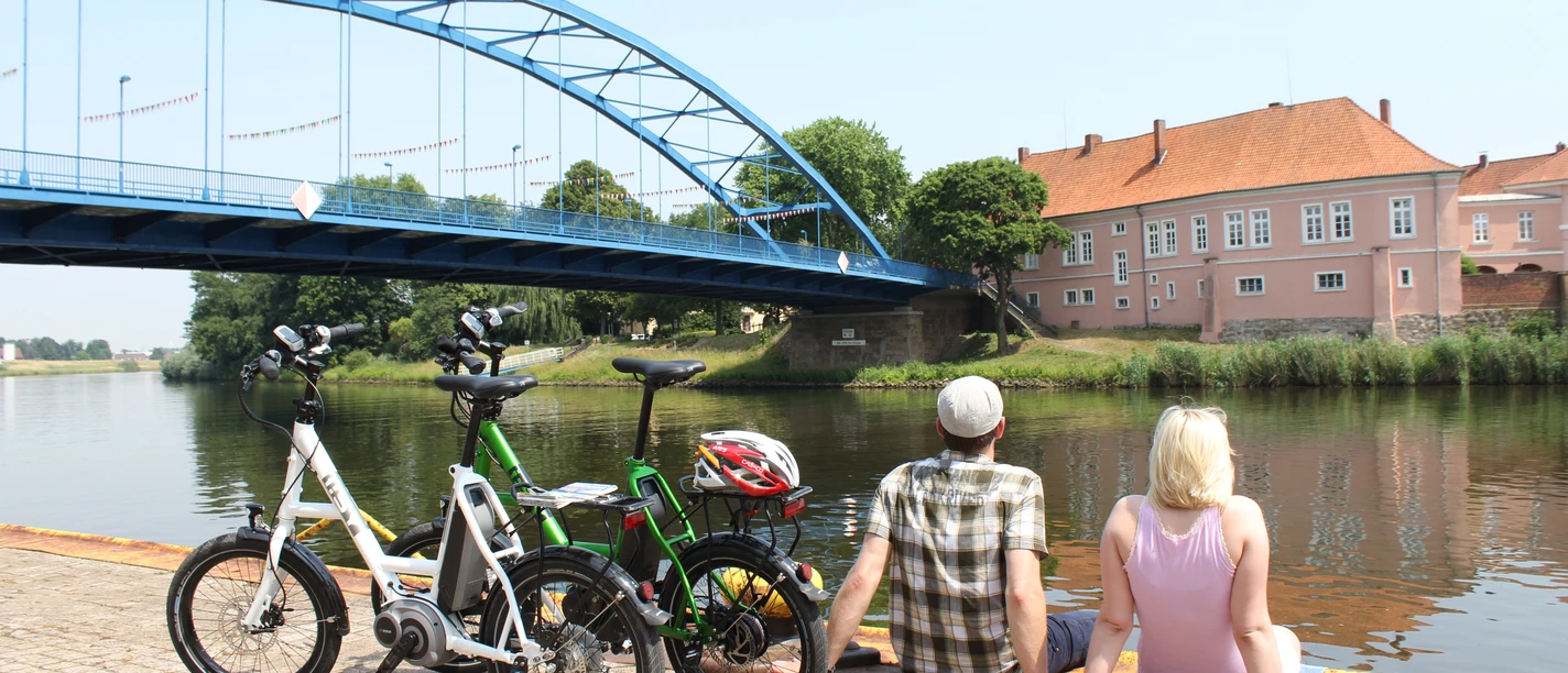 Grafenschloss zu Hoya A couple sits relaxed by the water, with a view of the historic Hoya Castle and a blue bridge.