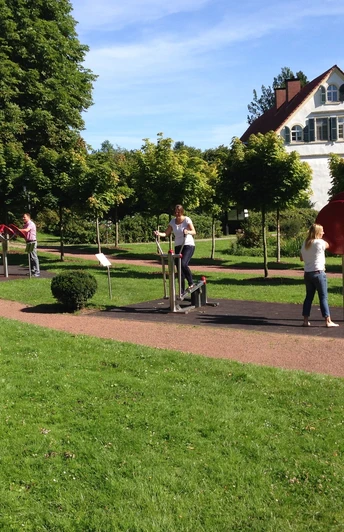 Garten der Generationen Menschen trainieren an Fitnessgeräten im Freien, umgeben von grüner Wiese und Bäumen, vor einem Haus.