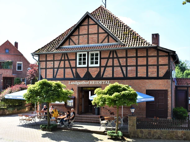 Landgasthof Heidetal Historisches Fachwerkhaus mit rotem Ziegeldach und Außenbewirtung unter blauen Sonnenschirmen. Vor dem Eingang sitzen Gäste an Holztischen im Schatten junger Bäume, während die Sonne die Backsteinfassade des Landgasthofs Heidetal freundlich beleuchtet.