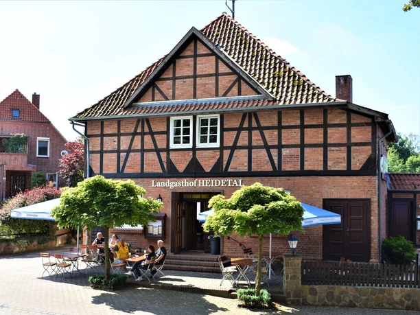 Landgasthof Heidetal Historisches Fachwerkhaus mit rotem Ziegeldach und Außenbewirtung unter blauen Sonnenschirmen. Vor dem Eingang sitzen Gäste an Holztischen im Schatten junger Bäume, während die Sonne die Backsteinfassade des Landgasthofs Heidetal freundlich beleuchtet.
