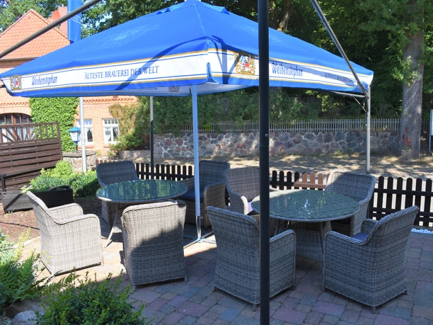 Außenterasse Außenterrasse mit mehreren runden Glastischen und geflochtenen Stühlen unter einem großen blauen Sonnenschirm. Die Sitzgruppe steht auf Pflastersteinen, umgeben von Pflanzen, Holzzaun und historischen Backsteingebäuden im Hintergrund.