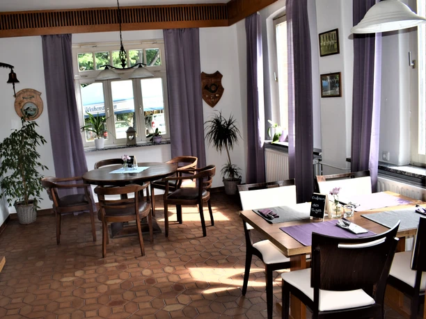 Gastraum Heller Gastraum mit großen Fenstern, lilafarbenen Vorhängen und Holzmöbeln. Zwei Tische sind eingedeckt, einer mit einer kleinen Tafel „Empfehlung des Tages“. Zimmerpflanzen und sanftes Tageslicht schaffen eine freundliche Atmosphäre in traditionellem Landhausstil.