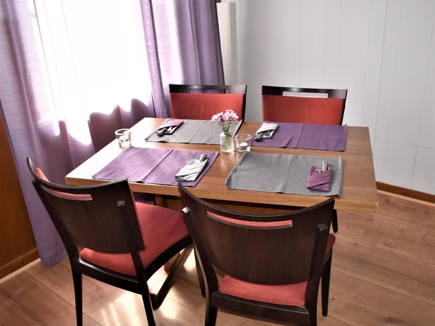 Gastraum Holztisch mit vier dunkelbraunen Stühlen, gedeckt mit grauen und lilafarbenen Platzsets, gefalteten Stoffservietten und Besteck. In der Mitte steht ein Glas mit frischen rosa Blüten. Sanftes Tageslicht fällt durch einen leicht geöffneten Vorhang in Fliederfarben auf den hellen Holzboden.