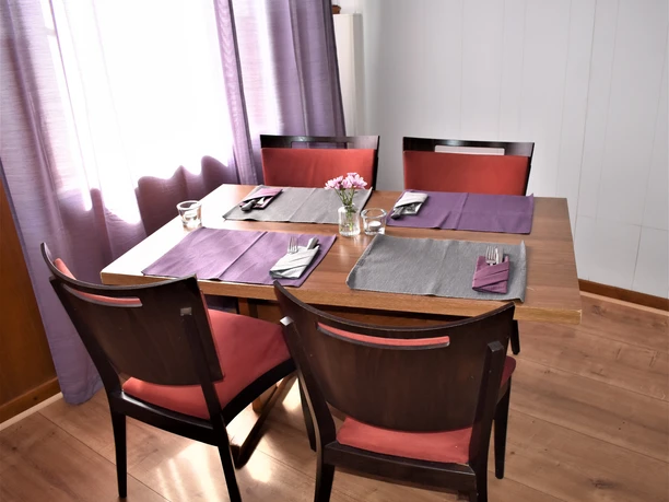 Gastraum Holztisch mit vier dunkelbraunen Stühlen, gedeckt mit grauen und lilafarbenen Platzsets, gefalteten Stoffservietten und Besteck. In der Mitte steht ein Glas mit frischen rosa Blüten. Sanftes Tageslicht fällt durch einen leicht geöffneten Vorhang in Fliederfarben auf den hellen Holzboden.