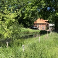 Klostermühle Heiligenberg Backsteinhaus mit rotem Dach, umgeben von üppigem Grün und einem Teich, an einem sonnigen Tag.