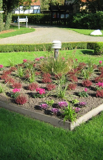 Kurwanderweg Ein gepflegtes, quadratisches Blumenbeet mit bunten Pflanzen in einem Park, umgeben von Rasen.