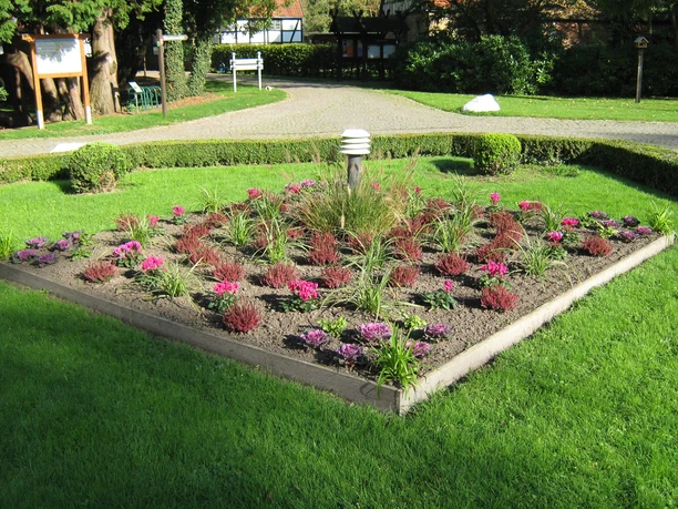 Kurwanderweg Ein gepflegtes, quadratisches Blumenbeet mit bunten Pflanzen in einem Park, umgeben von Rasen.
