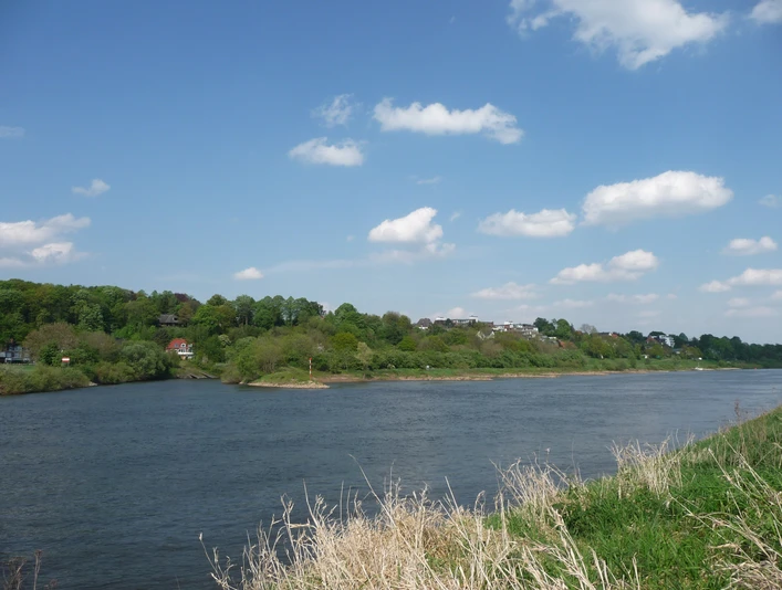 Grüne Hügel säumen das Ufer der friedlich fließenden Weser unter einem blauen, leicht bewölkten Himmel.