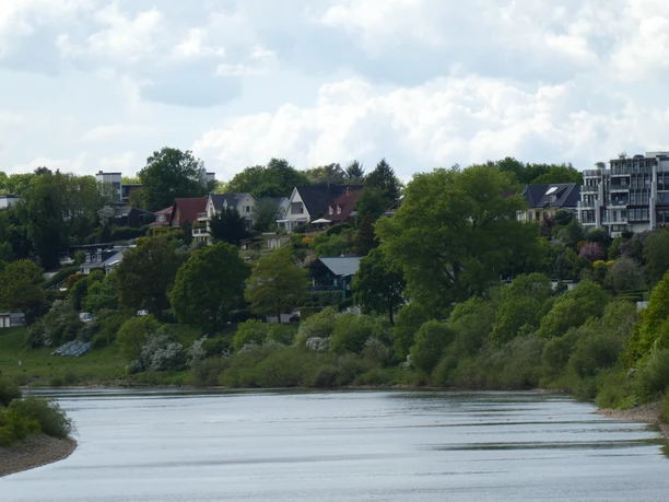 Badener Berge und Weser Flusslandschaft der Weser mit grünen Ufern und Wohnhäusern unter bewölktem Himmel.